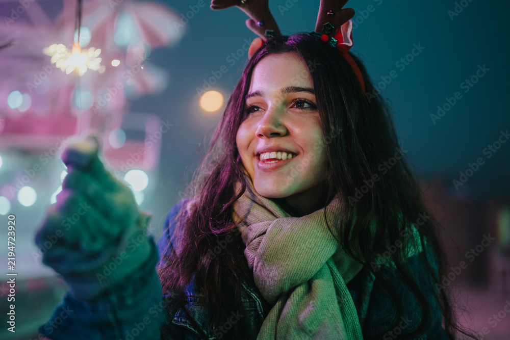 Obraz premium Young woman in a scarf and antler headband smiles while holding a lit sparkler. A festive winter market scene with colorful lights and friends enjoying the holiday evening.