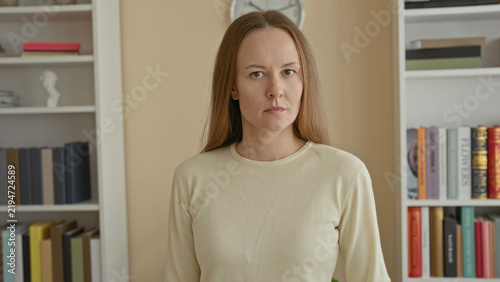 Obraz na plátně Woman standing with visible face looking at camera in studio framed by bookshelf and wall clock; calm contemplation