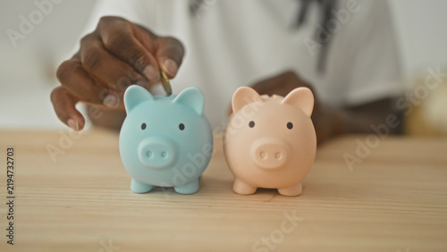 Konstfotografi Young african american man uses right hand to insert coin into pastel piggy bank pair on wooden table in studio; saving hope