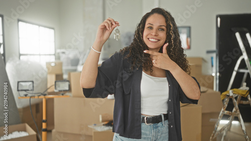 Fotografía Woman holding keys and pointing finger in apartment with moving boxes and packing materials; new home joy