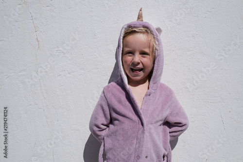 little girl wearing funny unicorn hooded bathrobe against white textured wall