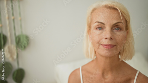 Foto Woman smiling face with exposed shoulder and freckles seated on bed in studio, soft pillow and plant visible; serenity maturity confidence