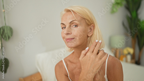 Quadro su tela Middle age blonde woman holding amber serum bottle, fingertip to cheek while applying skincare beside bed in a studio; self care calm wellness