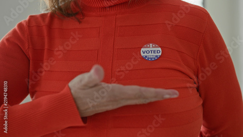 фотографія Woman points finger to voted sticker on her chest while wearing a red sweater at a polling station inside a building; civic pride