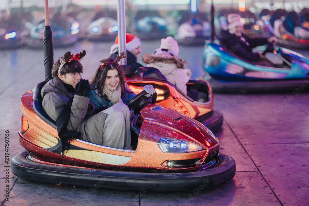 Obraz premium Young couple wearing reindeer antlers ride a glittering bumper car at a festive fairground. They smile and drive together among colorful lights and other bumper cars.