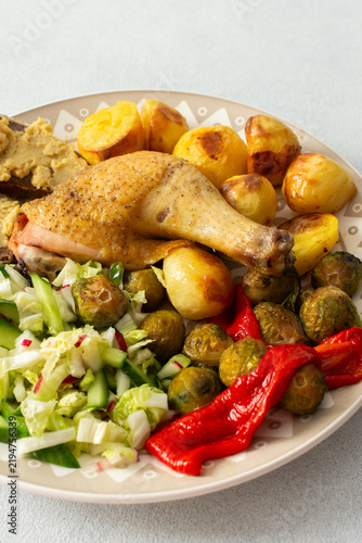 Foto A plate holds baked chicken leg, potatoes, fresh salad, roasted Brussels sprouts, and chickpea bread