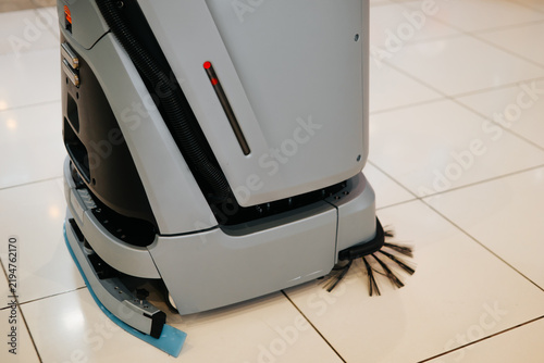 A robot floor cleaner is actively cleaning a tiled floor surface. The device features rotating brushes and is positioned at an angle as it works