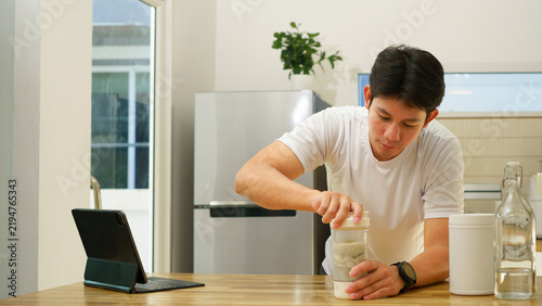 Foto Young Asian man preparing protein shake at home kitchen counter with tablet and