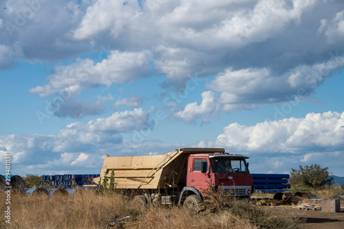 Cuadro en lienzo A weathered red dump truck sits parked beside stockpiled blue industrial pipes in an open, dry field under a vast, cloud-filled sky