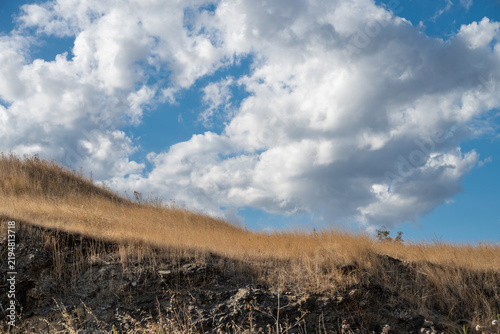 A golden, sunlit grassy hillside rises toward a vibrant blue sky filled with puffy white clouds, showcasing dry rural terrain and a partially shaded foreground slope.