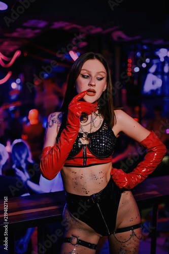 Female nightclub dancer poses in red gloves and black jeweled stage costume under colorful neon lights, creating a confident nightlife portrait for party and event promotion