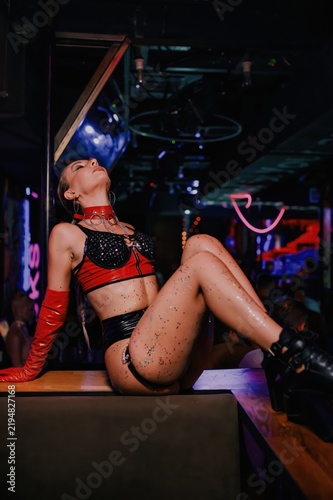 Nightclub performer reclines on a bar counter in red and black stagewear, glitter body makeup and platform boots, creating a dramatic nightlife image for dance and event promotion