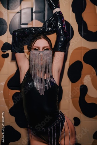 Female performer poses in black glossy stagewear, long gloves and sparkling rhinestone face fringe against an abstract wall, suited to nightlife, fashion and beauty advertising