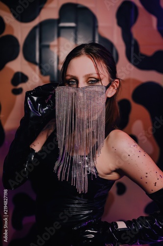 Close portrait of female performer wearing sparkling rhinestone fringe mask and glossy black glove against an abstract patterned wall, ideal for fashion, beauty and nightlife campaigns