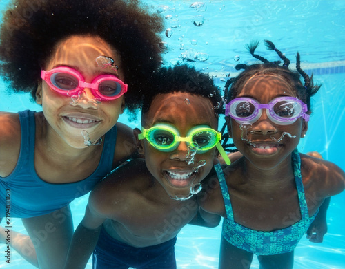 Group of cheerful children, girls and boy, are wearing swimming goggles and happily looking at the camera underwater in a blue swimming pool, with bubbles all around them, enjoying a relaxing spa.
