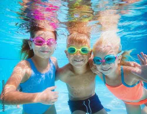 Group of cheerful children, girls and boy, are wearing swimming goggles and happily looking at the camera underwater in a blue swimming pool, with bubbles all around them, enjoying a relaxing spa.