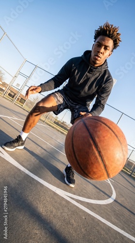 Vertical photo of a young Black man dribbling a basketball on an outdoor court. Low angle wide angle shot with perspective distortion and dutch tilt. Athletic streetball player training