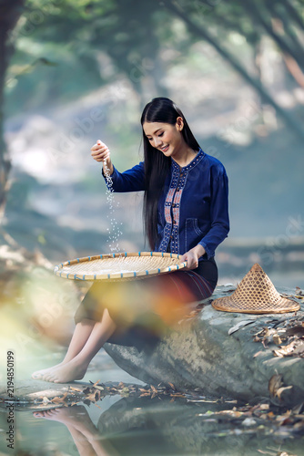 Smiling barefoot young woman in traditional Thai clothing sitting on a rock by a river winnowing rice (Fad Khao) using a bamboo tray (Kradong), Sakon Nakhon, Thailand