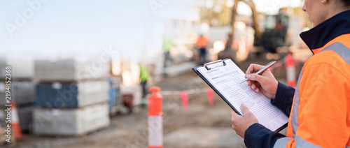Female construction site inspector in high-visibility safety vest holding clipboard and checking documents at an outdoor building development project with blurred industrial background.