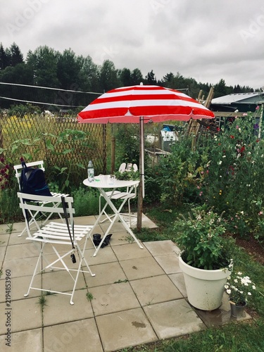 A paved garden patio features white folding furniture under a red and white striped parasol in a blooming garden.
