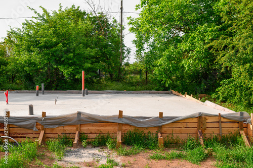 Fresh concrete slab foundation with wooden formwork and utility pipes surrounded by green trees in daylight, documenting residential construction and building development