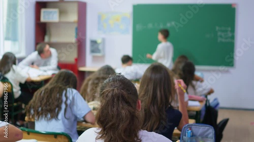 A group of young students sits at wooden desks in a bright classroom, while a classmate writes lesson notes on the large green chalkboard at the front of the room