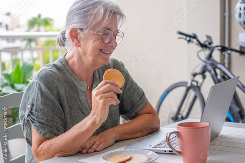 Принти на полотні Senior woman smiling and enjoying a cookie while engaging in a video call on her