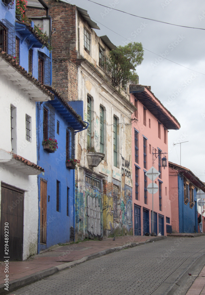 Fototapeta premium Colorful historic street in Bogotá’s La Candelaria, Colombia, featuring colonial buildings, traditional balconies, tiled roofs, graffiti, and vibrant facades.