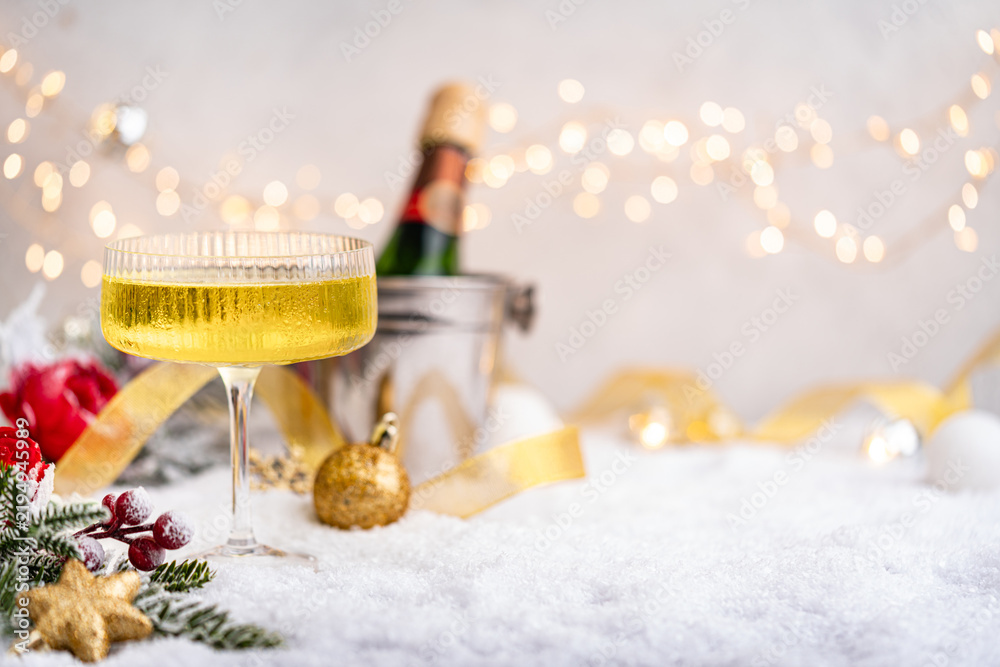 Obraz premium Elegant coupe glass filled with bubbly champagne sits on snow with a bottle, golden ribbon, and soft Christmas lights in a celebratory winter scene. Ideal for holiday greeting cards,