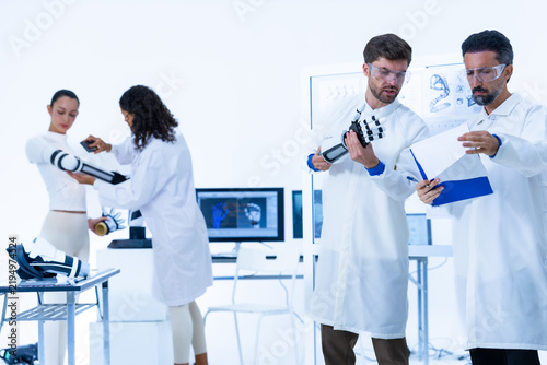 Obraz na plátně Two engineers review a clipboard while two researchers examine a pedestal roboti