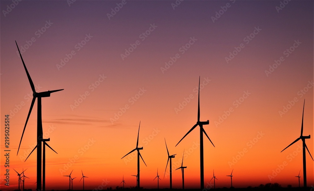 Obraz premium Vue panoramique d'un champ d'éoliennes dessinant des silhouettes verticales au coucher du soleil, sous un ciel orange mauve. Symbole d'une énergie renouvelable grâce à la force du vent.