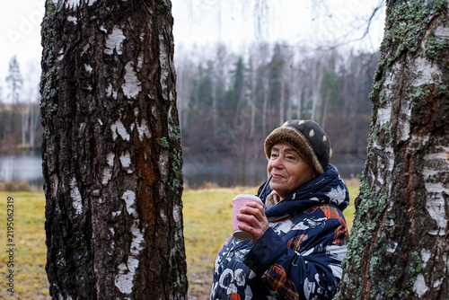 A happy smiling elderly woman enjoying a hot drink in a nature park.