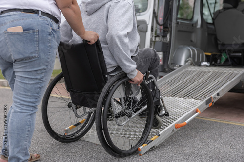 A person in a wheelchair receives help using a ramp to get into an adapted van. Personal assistant provides support in a parking area.