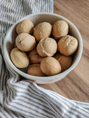 Whole walnuts in white ceramic bowl and striped kitchen cotton towel on wooden table.  Healthy eating, calories, diet, harvest. Shells of washed nuts are perfectly clean. Copy space. Close-up.