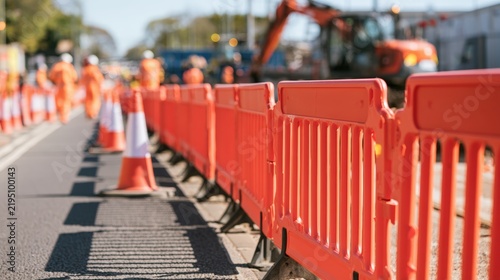 Construction site with orange safety barriers and traffic cones along a road, featuring workers in orange uniforms and machinery in the background, indicating ongoing roadwork and safety measures