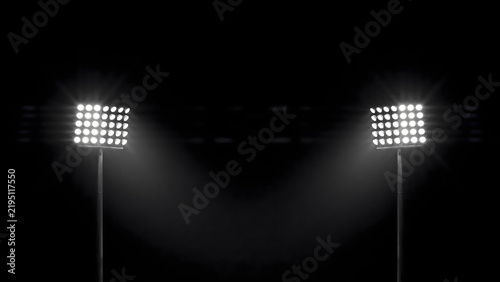 Two tall stadium floodlight towers glowing brightly against a dark night sky, illuminating the surrounding area with intense white light beams