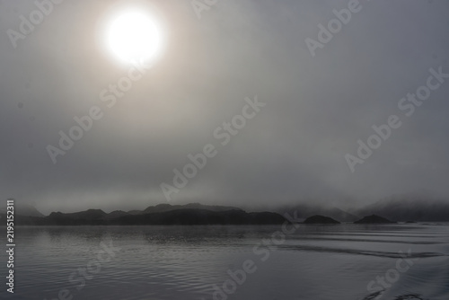 Glowing bright sun breaking through dense morning fog over calm sea waters with distant mountain silhouette, creating a serene landscape.
