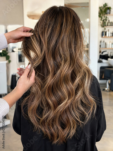 Long wavy hair with a caramel balayage seen from behind while the stylist lifts one strand