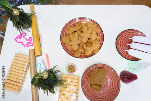 The kitchen table is covered with tools and ingredients for baking Christmas gingerbread cookies. Dough, reindeer shapes, and sprinkles are arranged amidst holiday decorations.