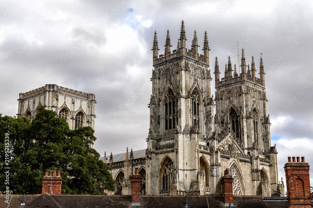 Fototapeta premium York Minster rises above historic rooftops beneath dramatic cloudy skies in York, England, showcasing the Gothic architecture and ornate towers of the medieval cathedral