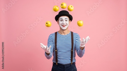 Commercial performance stock photo of a male mime in a striped shirt and bowler hat juggling bright yellow apples against a solid pink background.