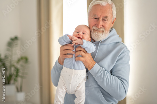 Fotografía Elderly man holds baby in living room with green plant behind, grandfather meeting grandson first months, feeling everyone recognises