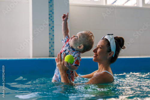 A mother lifts her baby above pool water as the infant holds a green ball and points one finger upward