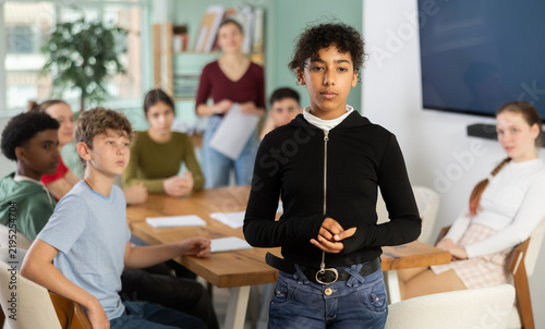 Fotografie Sad teenage girl standing with her back to classmates sitting at table in classr