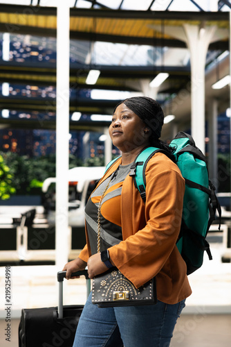 Фотография Woman passenger carries a backpack and pulls trolley luggage across a bus station, moving through the terminal on an intercity journey toward her planned route with a relaxed weekday travel mood