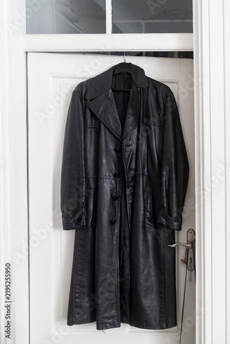 Long black leather coat hanging on a hanger against a white wooden door.
