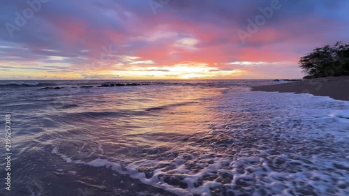 Waves hit the shore as the sun rises, casting colors in the sky at a beach. This scene shows nature's beauty at dawn.