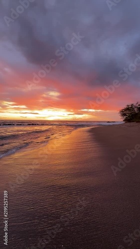 The sun sets over the ocean as waves wash onto the sandy beach. The sky shows bright colors in the evening light near a shoreline.