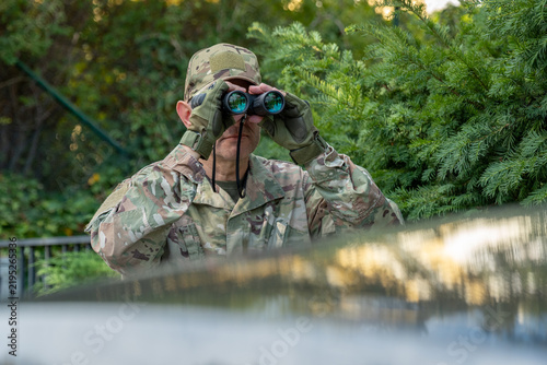 Konstfotografi Intelligence scout monitors a distant target with optics while remaining concealed near a vehicle and dense vegetation