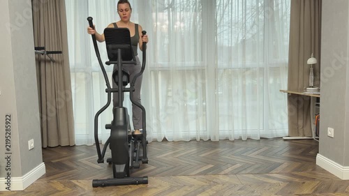Woman steps forward on elliptical machine synchronizing arm and leg movement. Effective home cardio session in modern living room.